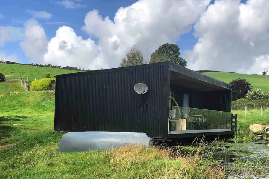 Glamping Pods North Wales view of black camping pod with pond in front, boat and the balcony with chairs